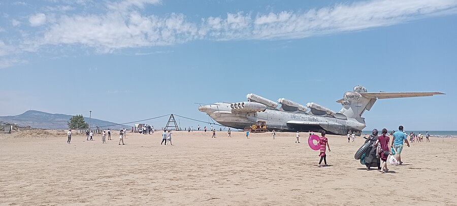 The Lun ekranoplan MD-160 beached on a pebble shore: long dark hull, eight turbofans forward, missile containers along the spine, tall fin at the stern.